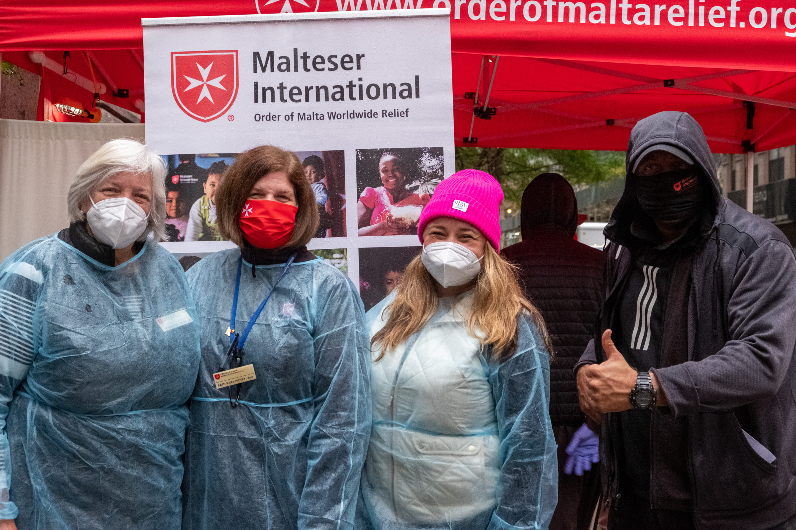 Three nurses and a beneficiary stand in front of a Malteser International Americas sign.